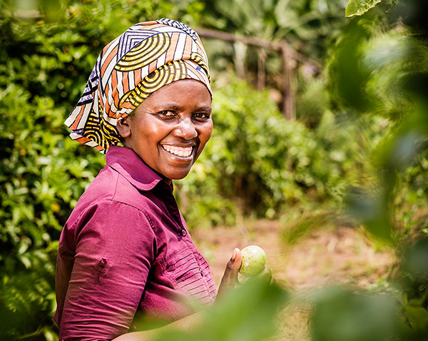 En leende kvinna står i en prunkande miljö med en frukt i handen.